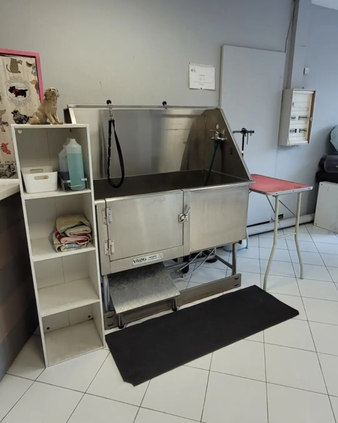 Zone de lavage libre service pour animaux de compagnie avec une grande baignoire en acier, des serviettes rangées sur une étagère et une table de toilettage rose à côté.
